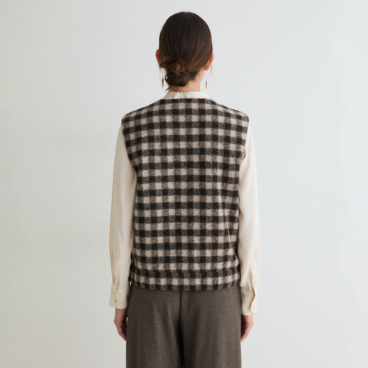 A model shown from the back wearing a sleeveless black and beige checkered vest over a cream long-sleeve blouse and brown trousers, set against a plain light gray background.