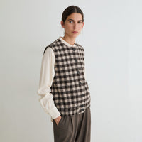 A woman modeling a sleeveless black and beige checkered vest over a cream long-sleeve blouse and brown trousers, standing against a plain light gray background.