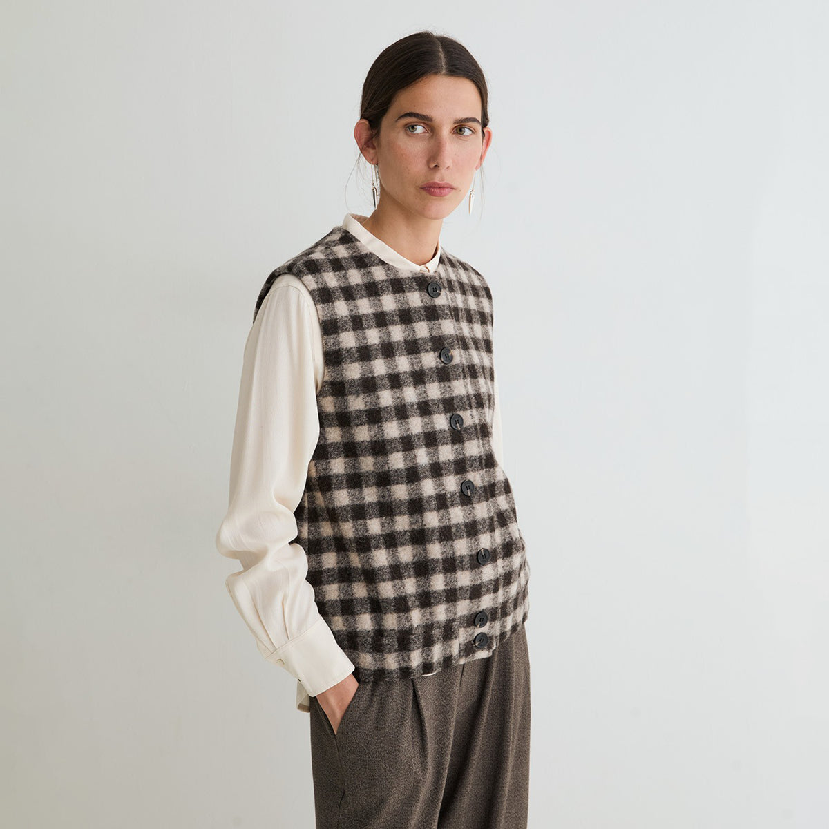 A woman modeling a sleeveless black and beige checkered vest over a cream long-sleeve blouse and brown trousers, standing against a plain light gray background.