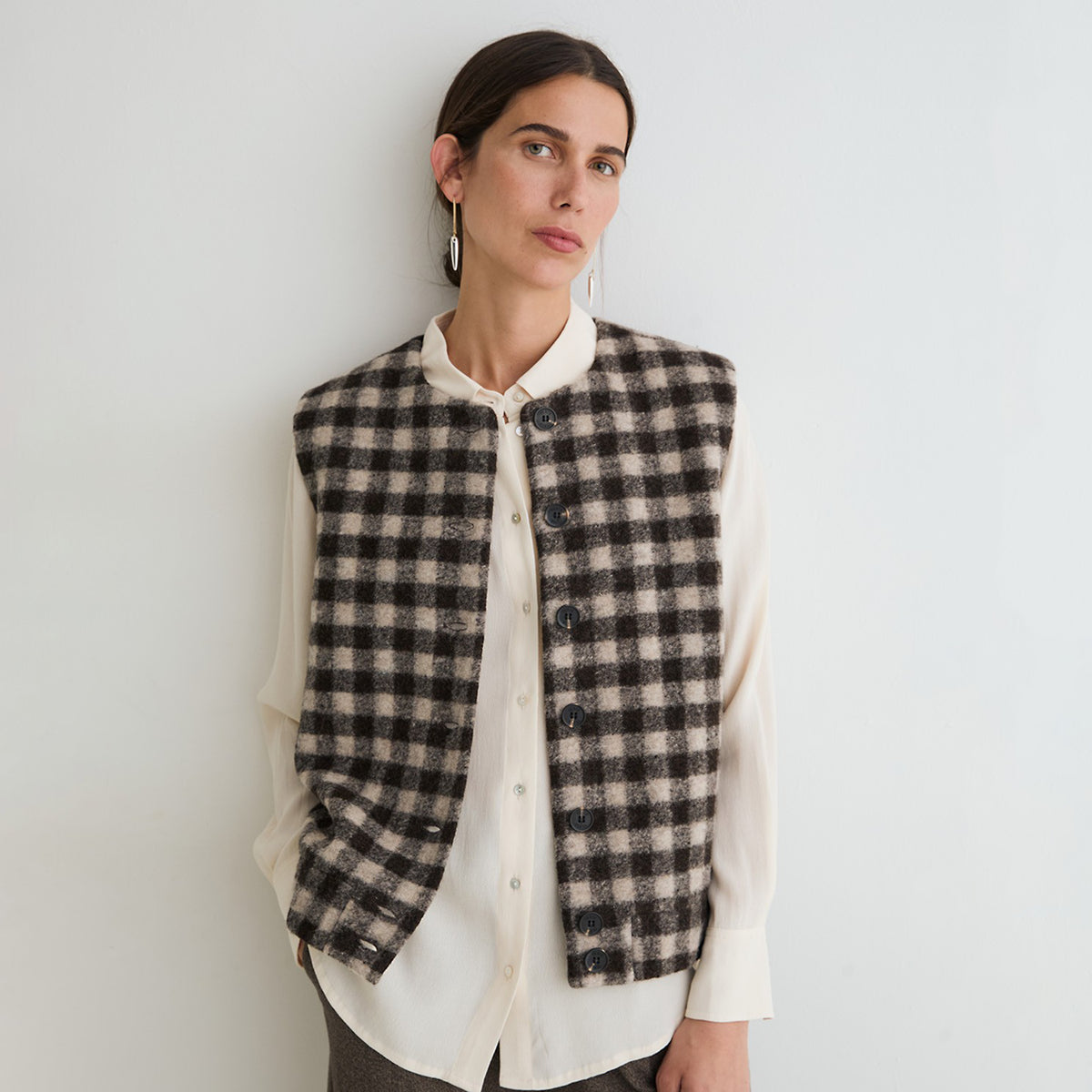 A woman wearing a black and beige checkered button-front vest over a cream button-up shirt, standing against a plain light gray wall.