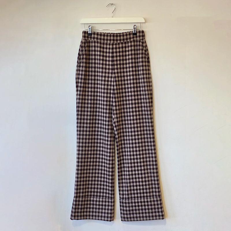 A pair of wide-leg cropped pants in a brown and beige gingham check pattern, hanging from a white plastic hanger against a plain off-white wall.