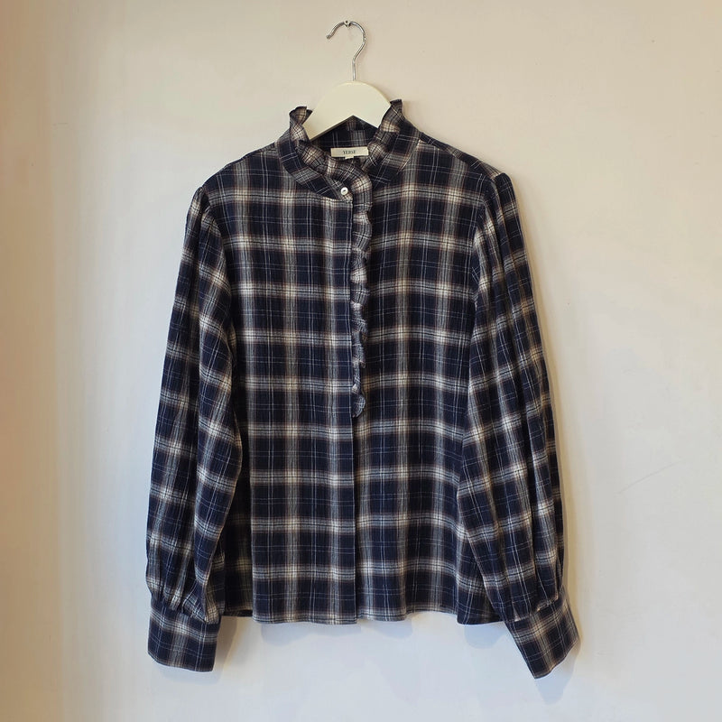 A long-sleeve plaid button-up shirt with a ruffled mock neck and ruffle trim down the front placket, hanging on a white hanger against a plain off-white wall.