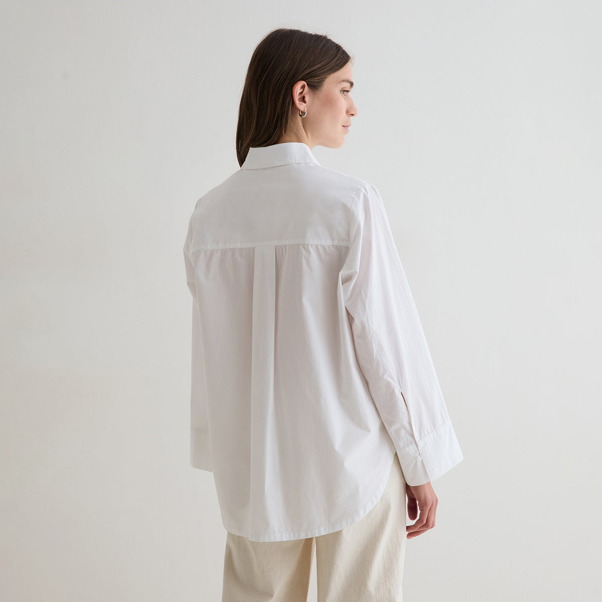 A woman shown from the back wearing an oversized white button-up shirt and light beige trousers, photographed against a plain off-white studio background.
