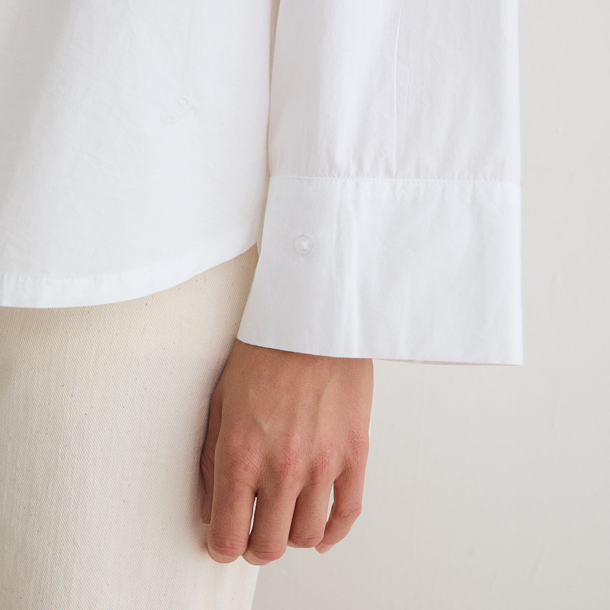 Close-up of a person wearing a white long-sleeve button-up shirt with a wide cuff and a light beige textured skirt or pants, with the hand resting near the fabric.