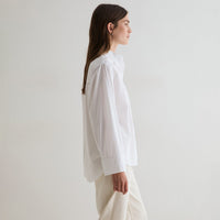 A woman in profile wearing a loose white long-sleeve blouse and light beige trousers, standing against a plain off-white studio background.