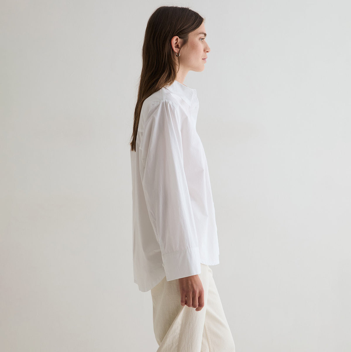 A woman in profile wearing a loose white long-sleeve blouse and light beige trousers, standing against a plain off-white studio background.