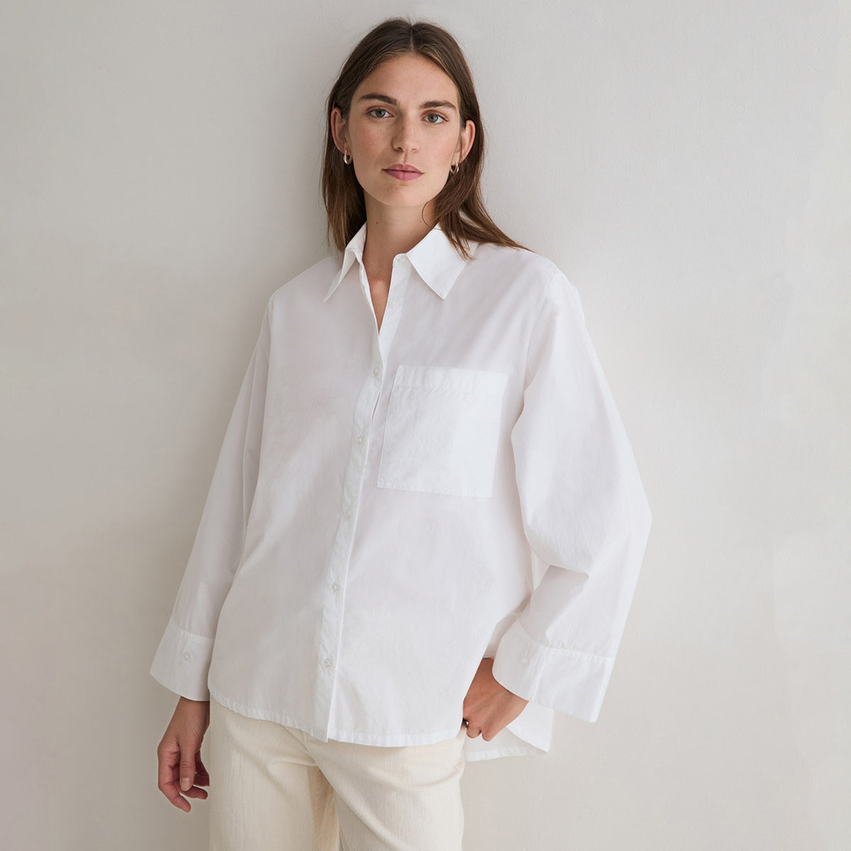 A woman wearing an oversized white long-sleeve button-up shirt with a chest pocket, paired with cream pants, standing against a plain light gray wall.