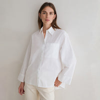 A woman wearing an oversized white long-sleeve button-up shirt with a chest pocket, paired with cream pants, standing against a plain light gray wall.