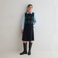 A woman stands against a plain light gray wall wearing a light blue long-sleeve collared shirt, a dark navy argyle sweater vest, a dark navy pleated skirt, and black knee-high boots.