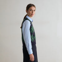 A woman in profile wearing a navy and green argyle sweater vest over a light blue collared shirt and a dark skirt, photographed against a plain light gray studio background.