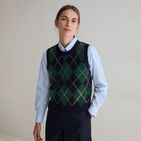 A woman wearing a navy and green argyle knit sweater vest over a light blue collared shirt and dark trousers, standing against a plain light gray studio background.