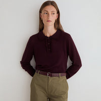 A woman wearing a dark burgundy long-sleeve knit top with a ruffled collar and button placket, paired with olive green trousers and a matching burgundy belt, standing against a plain off-white wall.