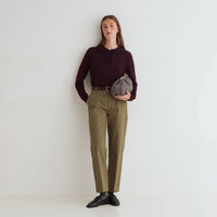 A woman stands against a plain white wall wearing a dark burgundy long-sleeve polo shirt, olive green trousers, a black belt, and black leather shoes. She is holding a small gray and black patterned clutch bag.