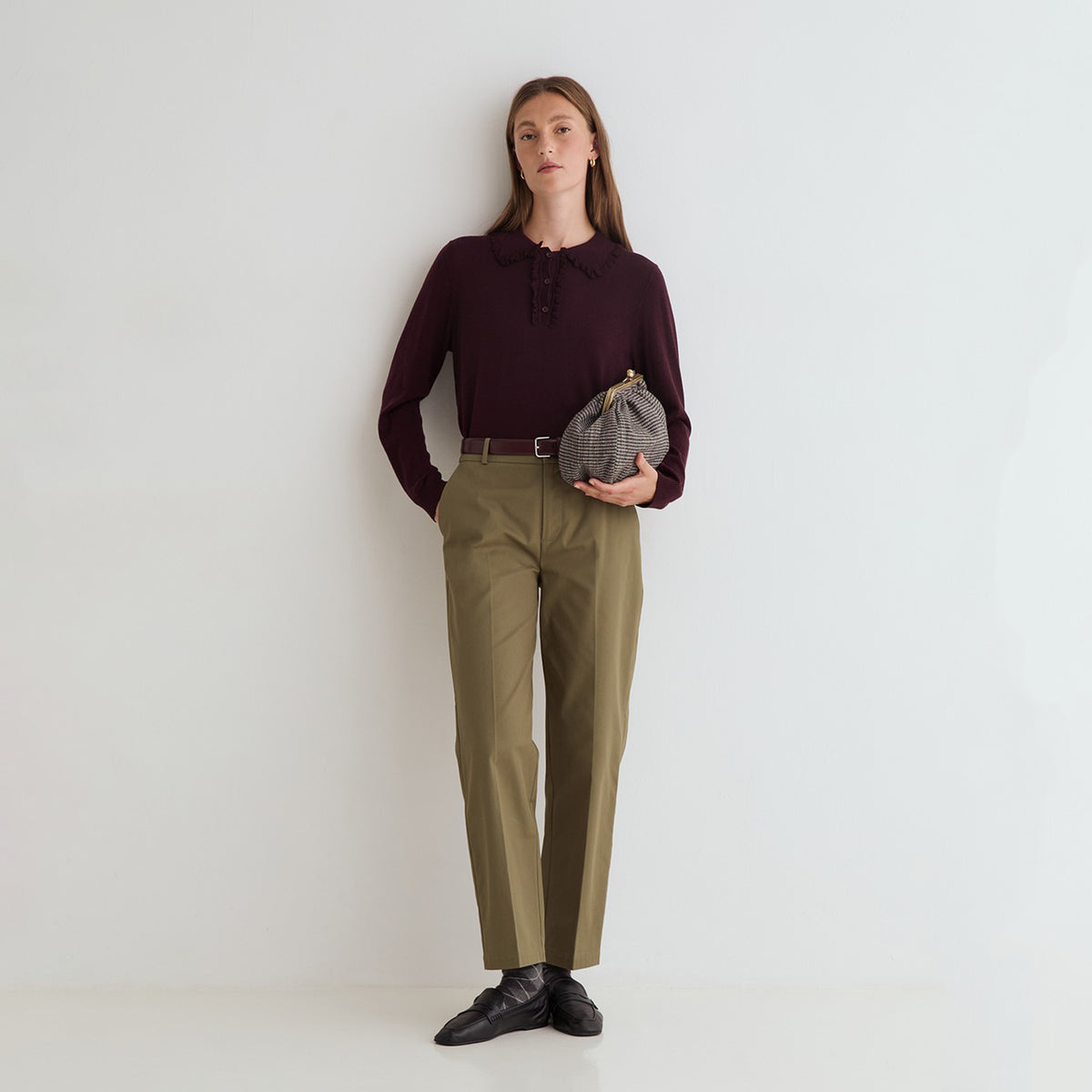 A woman stands against a plain white wall wearing a dark burgundy long-sleeve polo shirt, olive green trousers, a black belt, and black leather shoes. She is holding a small gray and black patterned clutch bag.