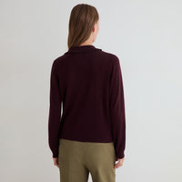 A woman viewed from the back wearing a dark burgundy knit sweater and olive green pants against a plain light gray studio background.