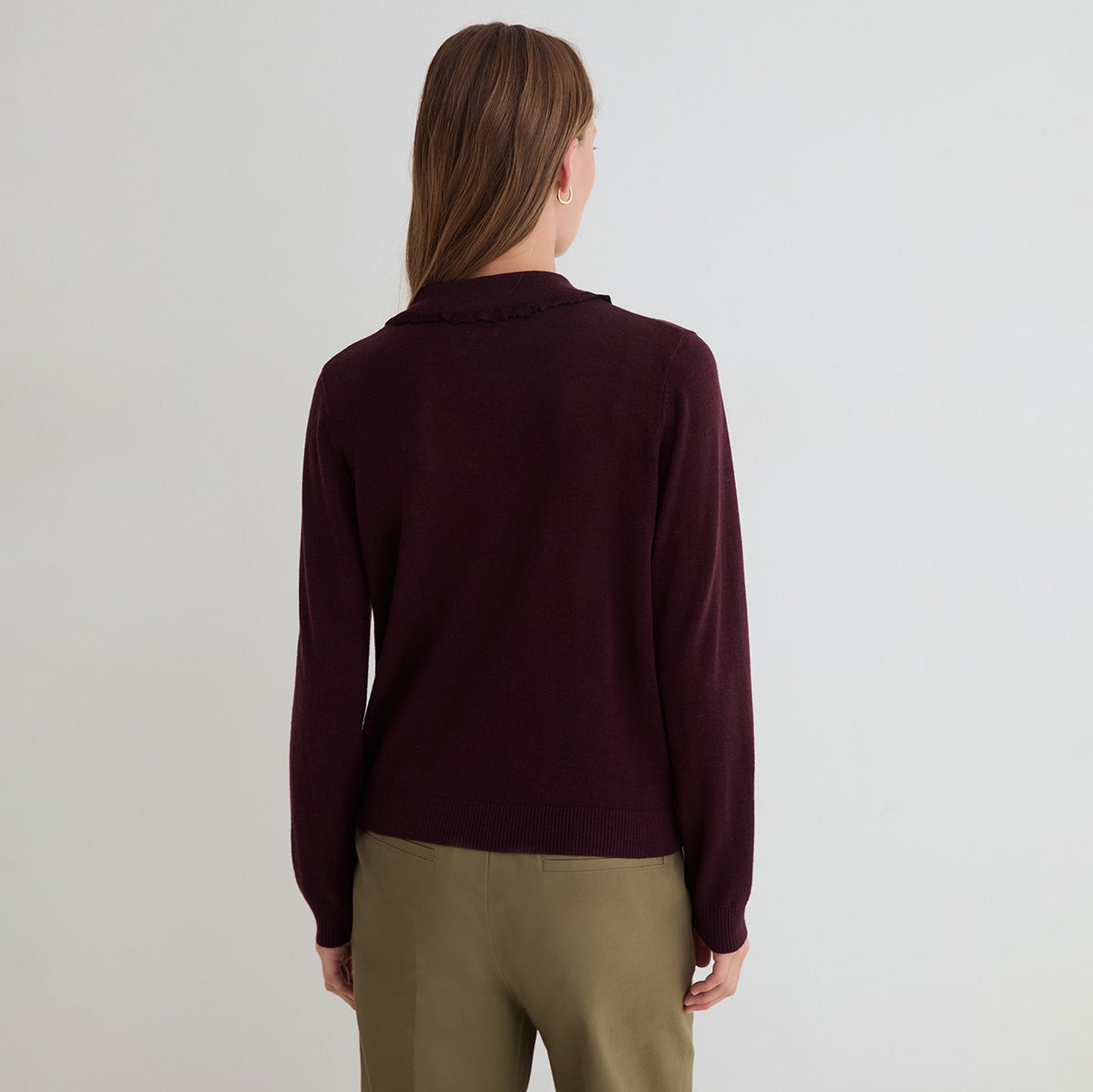 A woman viewed from the back wearing a dark burgundy knit sweater and olive green pants against a plain light gray studio background.