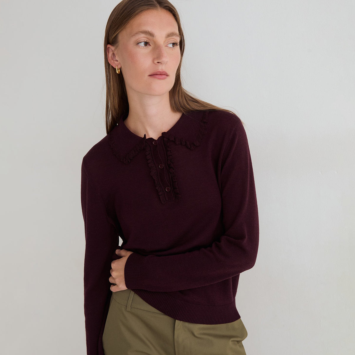 A woman wearing a dark burgundy long-sleeve knit sweater with a ruffled collar and button placket, paired with olive green pants, photographed against a neutral light gray background.