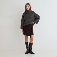 A model stands against a plain white studio background wearing a dark gray oversized turtleneck sweater, a dark brown wrap-style mini skirt with fringe, and black knee-high leather boots.