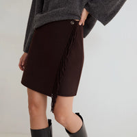 A person wearing a dark brown wrap-style mini skirt with fringe trim and a gray knit sweater, paired with black boots, against a plain light background.