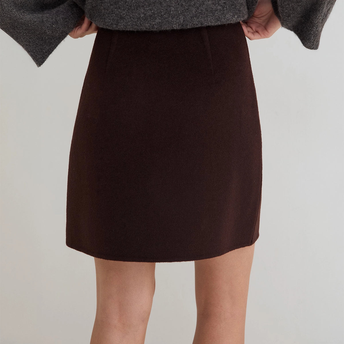 A dark brown wool A-line mini skirt worn by a model, paired with a gray knit sweater, shown from the waist down against a light gray background.