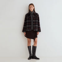 A model stands against a plain white studio wall wearing a dark plaid button-front jacket over a gray turtleneck, a dark brown fringe mini skirt, and black knee-high boots.