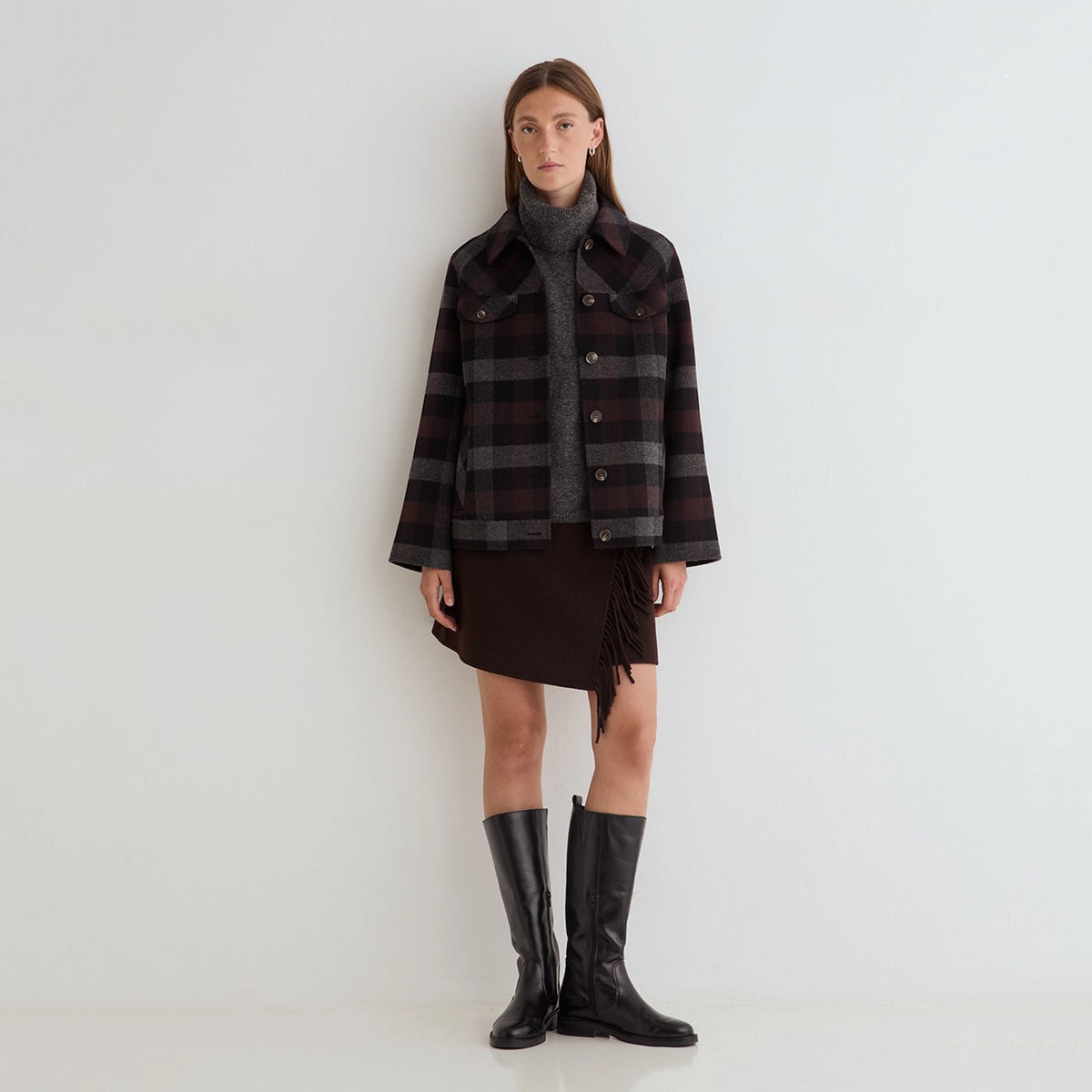 A model stands against a plain white studio wall wearing a dark plaid button-front jacket over a gray turtleneck, a dark brown fringe mini skirt, and black knee-high boots.
