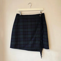 A dark navy and green plaid wrap skirt with fringe detailing, hanging from a white plastic hanger against a plain beige wall.