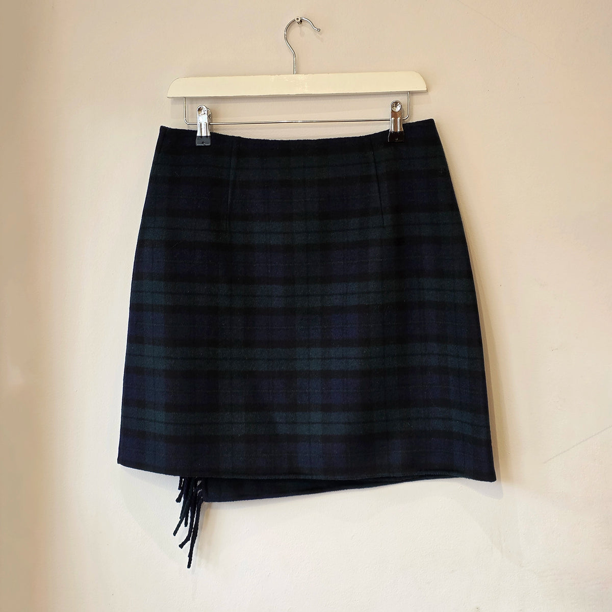 A dark navy and green plaid mini skirt with fringe detailing at the hem, hanging from a white wooden hanger against a plain beige wall.