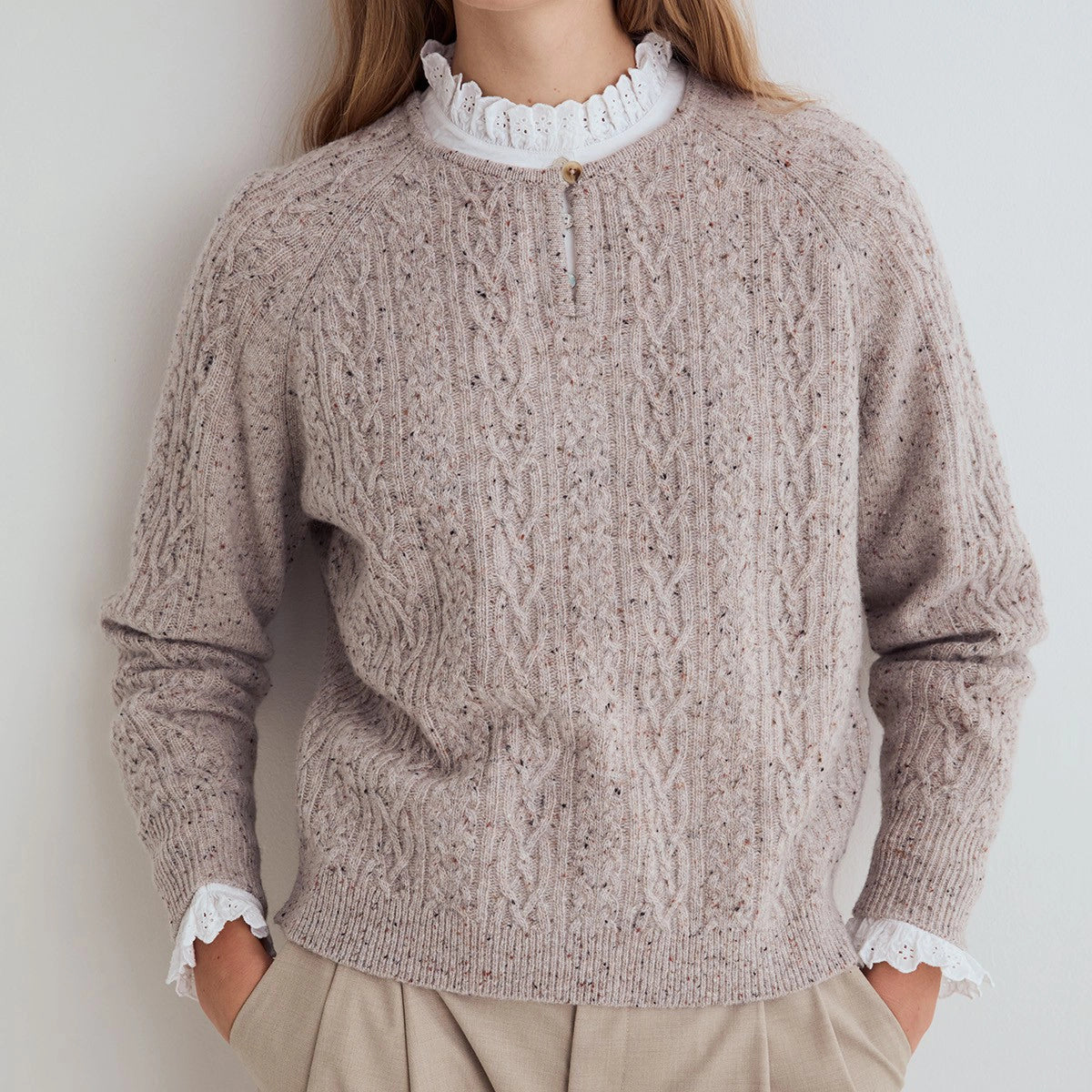 A woman wearing a taupe cable-knit sweater with white lace trim at the mock neck and cuffs, paired with beige trousers, photographed against a neutral light gray background.