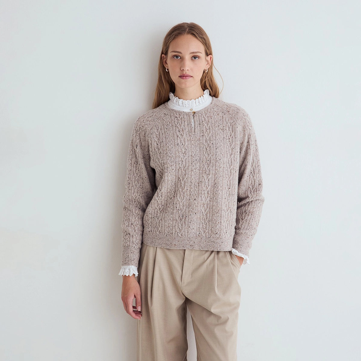 A woman stands against a plain light gray wall wearing a beige cable-knit sweater with white ruffled trim at the mock neck and cuffs, paired with beige trousers.
