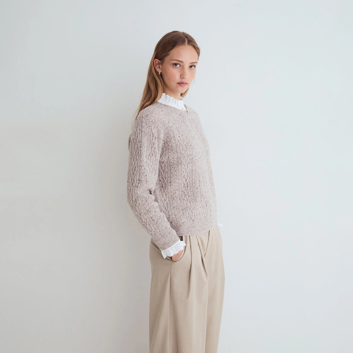 A woman stands in profile wearing a textured light beige knit sweater over a white ruffled mock neck blouse, paired with beige wide-leg trousers, against a plain light gray studio background.