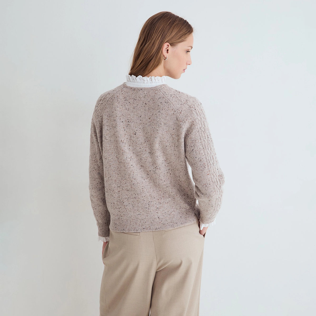 A woman shown from the back wearing a light taupe speckled knit sweater with a white ruffled collar, paired with beige trousers, against a plain light gray background.