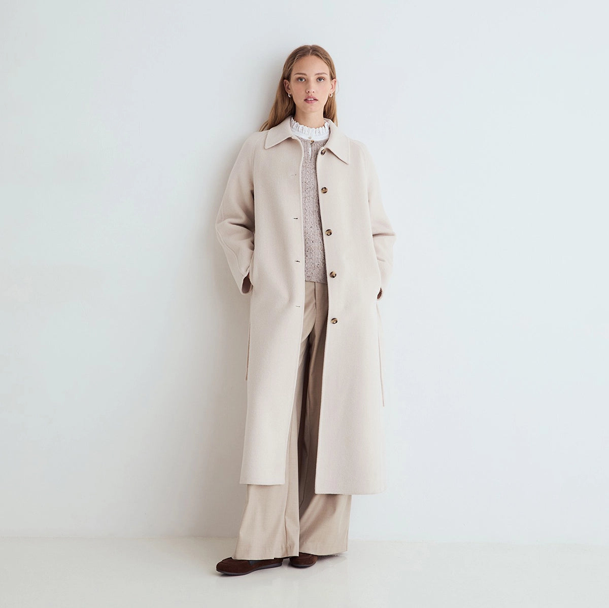A woman stands against a plain white wall wearing a long light beige overcoat over a textured knit top and wide-leg trousers.