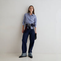 A woman stands against a plain light gray studio wall wearing a light blue and white striped button-up shirt, dark blue patchwork trousers, and dark pointed flats. She is holding a small dark bag.