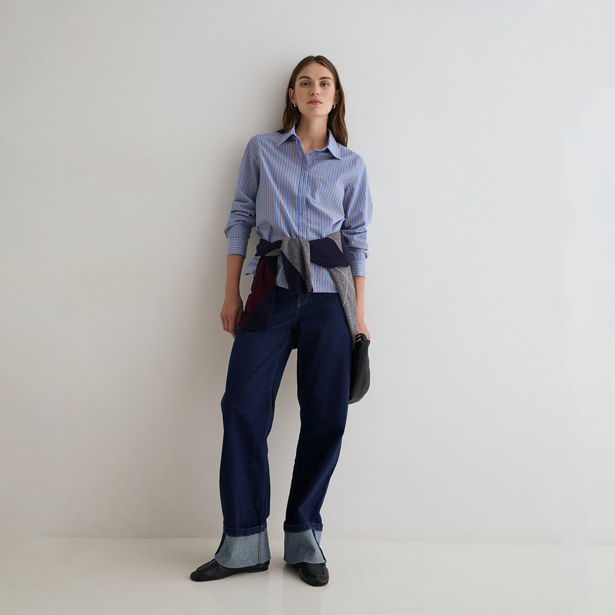 A woman stands against a plain light gray studio wall wearing a light blue and white striped button-up shirt, dark blue patchwork trousers, and dark pointed flats. She is holding a small dark bag.