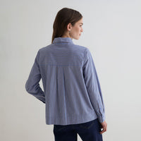 A woman shown from the back wearing a blue striped button-up shirt with thin red and white vertical lines, paired with dark blue jeans, against a plain light gray studio background.