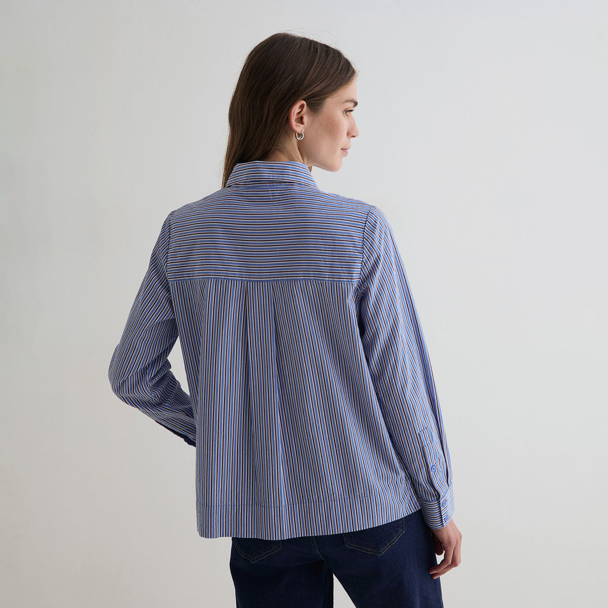 A woman shown from the back wearing a blue striped button-up shirt with thin red and white vertical lines, paired with dark blue jeans, against a plain light gray studio background.
