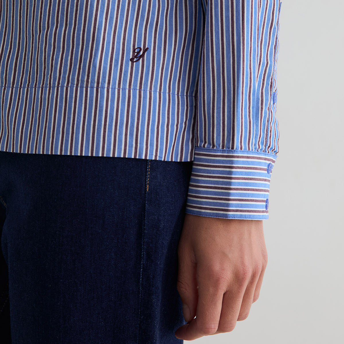Close-up of a person wearing a blue, white, and brown vertically striped long-sleeve shirt with a dark navy embroidered logo on the chest, paired with dark blue jeans.