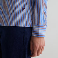 Close-up of a person wearing a blue, white, and brown vertically striped long-sleeve shirt with a dark navy embroidered logo on the chest, paired with dark blue jeans.