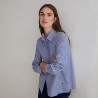 A woman wearing a blue and white vertically striped button-down shirt, standing with her arms crossed against a plain light gray wall.