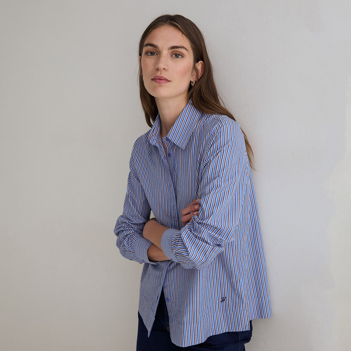 A woman wearing a blue and white vertically striped button-down shirt, standing with her arms crossed against a plain light gray wall.