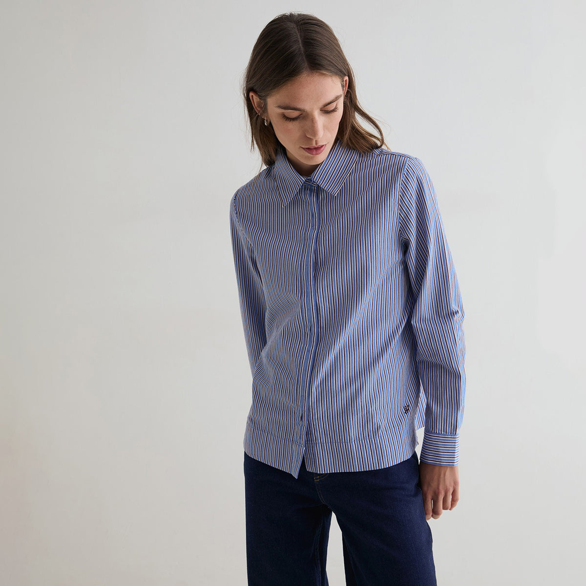 A woman wearing a blue and white striped button-down shirt with dark navy pants, standing against a plain light gray background.