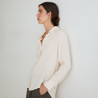 A woman in profile wearing a long-sleeve cream-colored button-up blouse and dark gray trousers, standing against a plain light gray wall.
