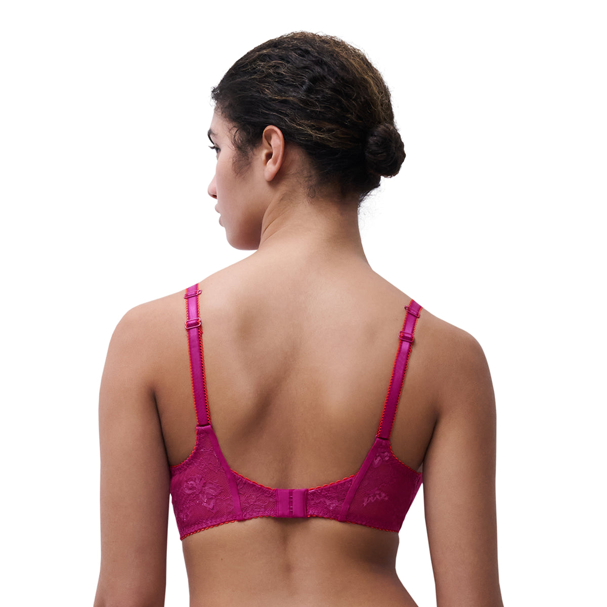 A woman shown from the back wearing a bright magenta lace bra with adjustable shoulder straps and a center gore, photographed against a plain white studio background.