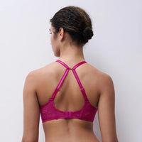 A woman shown from the back wearing a bright pink lace bra with crisscross shoulder straps, against a plain light gray background.