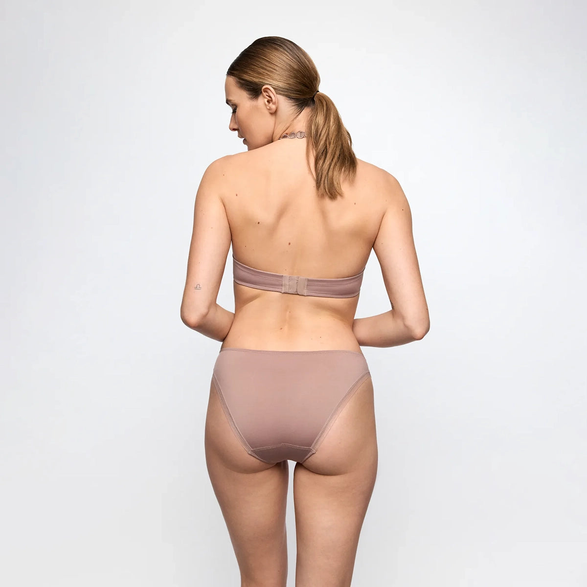 A woman shown from the back wearing a matching two-piece underwear set in a muted dusty rose or mauve color, photographed against a clean light gray studio background.