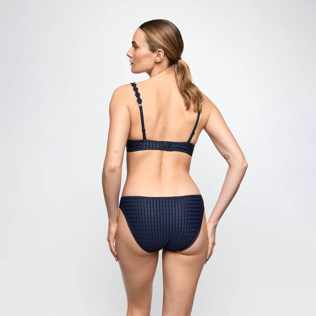 A woman shown from the back wearing a dark navy textured bikini set against a light gray studio background.