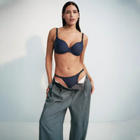 A woman modeling a dark navy blue textured two-piece lingerie set consisting of a bra and matching panty, styled with loose gray trousers against a light gray studio background.