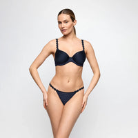 A woman modeling a two-piece bikini set in dark navy blue against a light gray studio background. The bikini features textured, scalloped straps on the bra and matching patterned bottoms.