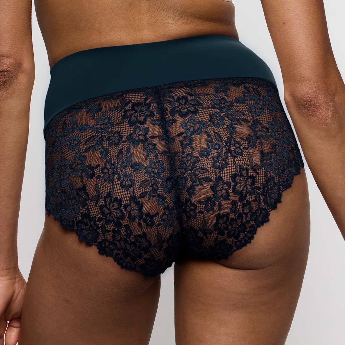 Rear view of a person wearing dark navy lace panties with a solid navy waistband, shown against a light gray studio background.